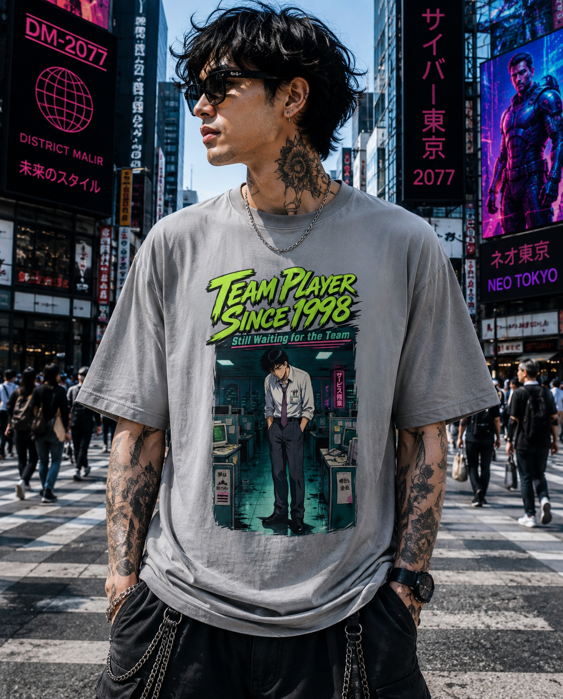 A man wearing a light gray graphic t-shirt with a retro comic-style print reading 'Team Player Since 1998' stands in a busy Tokyo street crosswalk. The background features tall city buildings with Japanese signage and neon lights.